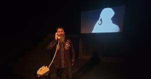 Comedian and filmmaker Jon Schnitzer with a long beard stands holding a vintage 1980s telephone to his ear. Behind him, a projection displays a silhouette of a person on a phone talking to him. Something Spooky is a comedy horror show that makes you scream and laugh.