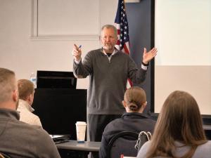 Jon Fehlman, founder of Thatcher Lewis, speaking with hands raised mid-presentation in front of an American flag during a leadership training workshop.