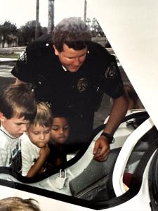 Young Officer Jon Fehlman in dark patrol uniform leans over an open police vehicle, showing three young children the interior during a community engagement event with palm trees visible in the background.