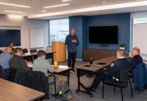 Jon Fehlman, founder of Thatcher Lewis, stands at the front of a modern training room in a navy quarter-zip pullover delivering a command leadership session to seated law enforcement officers, with a uniformed officer visible in the foreground.
