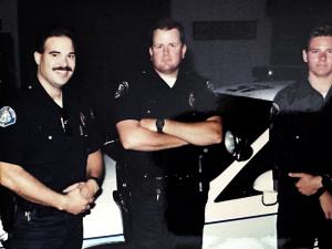Three uniformed police officers stand in a station garage in front of patrol cruisers, with a young Jon Fehlman at center wearing a duty watch and arms crossed, and a fellow officer with a mustache smiling at left.