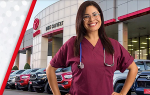 A nurse stands in front of Mike Calvert Toyota, a Houston car dealership. There is a row of Toyota trucks behind her, but in front of the dealership and the color of her scrubs are maroon as well as she has a stethoscope around her neck.