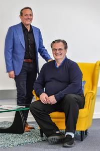 GT Clocks President Colin Leatherbarrow stands beside Stephen Sardi, VP Customer Success, who is seated in a yellow chair.