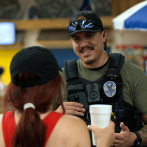 Police officer wearing a body camera and uniform talks to a woman with red hair and a black cap in an indoor setting.