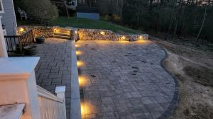 Custom paver patio installation by Backyard Experts in Massachusetts, featuring herringbone-pattern pavers with curved border, a natural fieldstone retaining wall with built-in steps, and integrated landscape lighting at dusk.