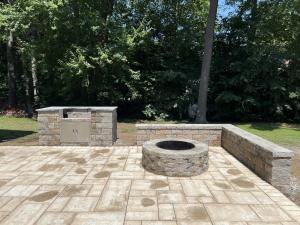 Complete outdoor living space by Backyard Experts in Massachusetts, featuring a multi-tone paver patio, custom stone outdoor kitchen with stainless steel grill, round stone fire pit, and L-shaped seat wall.