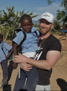 Gabe Butterfield smiles while holding a young child at a Vacation Bible School site during a mission trip to Africa, wearing a white God Tap hat outdoors in a rural community setting.