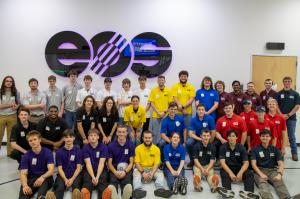 A large group of students wearing different colored shirts stand together in front of an EOS wall sign during an additive manufacturing competition event.