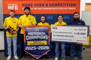 A group of students stands in front of a blue and orange Project MFG backdrop while holding a National Champions banner and a ceremonial $100,000 check.