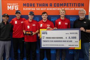 A group of students stands in front of a blue and orange Project MFG backdrop while holding a ceremonial $2,500 check.
