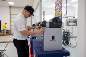 A student wearing a white shirt and black hat stands infront of 3D printer in an additive manufacturing shop