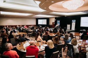 Zach Anner presents at the UCP Annual Conference. The conference room is organized with several tables all facing a stage with a black backdrop and screens on either side. Attendees are facing the stage and engaged with the presenter.