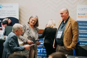 Three people are having a conversation in an exhibitor hall. There are branded booth backdrops behind them that include information about various disability-related services.