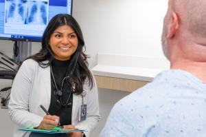 A KCU student wearing a white coat and stethoscope speaks with a seated patient in an exam room, taking notes on a clipboard; chest X-ray images are displayed on a monitor in the background.