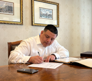 A professional photograph of Marcelo Umanzor Blanco wearing a white dress shirt, seated at a wooden desk and signing legal documents in a well-appointed office.