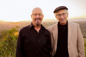 two gentlemen musicians standing in a seaside meadow, one in a hat