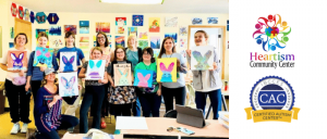 A group of people in a colorful art classroom holding up butterfly paintings, with Heartism Community Center and CAC logos beside the image.