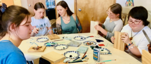 A group of children and an adult sitting around a table crafting with wooden boards, paint, and brushes in a classroom setting.