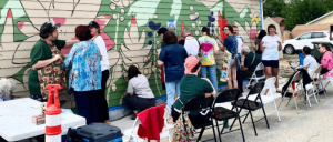 A group of people of various ages paint a colorful mural on the side of a building, with tables and chairs set up nearby.