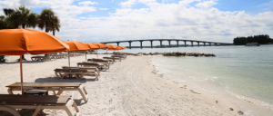 Beach lined with empty lounge chairs and orange umbrellas, palm trees, calm water, and a bridge under a partly cloudy sky.
