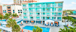Hotel building with balconies, a swimming pool, lounge chairs, and palm trees under a bright blue sky.