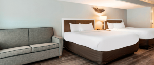 Hotel room with two queen beds, brown headboards, white bedding, a gray sofa, wooden floor, and a lamp on the nightstand.