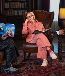 Author Emma Straub sits in a high-backed leather library chair, looking at a copy of Miss Piggy's Guide to Life held by Chris Collins.