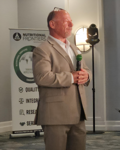 Jamie Dorley, founder of Nutritional Frontiers and co-host of The Holistic Truth Podcast, speaks at an event while holding a microphone in front of company signage.