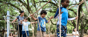 Children navigating a rope bridge in an outdoor park surrounded by trees, with an adult observing nearby.