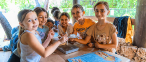 A group of six smiling children sit around a picnic table in a park, engaging in a crafting activity with colorful materials. The scene conveys joy and collaboration.