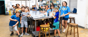 A group of children and adults gathered around a table in a classroom or workshop setting, smiling and posing for the photo.