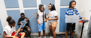 Five young musicians in a studio jam session. Two play guitars, two sing into a microphone, and one operates a keyboard and laptop. The atmosphere is lively and collaborative.