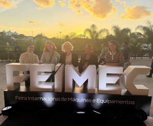 Cinco mujeres posan sonrientes detrás de una gran instalación iluminada con las letras “FEIMEC”, en un espacio exterior de la Feria Internacional de Máquinas y Equipos. Al fondo, el cielo muestra tonos dorados del atardecer, con nubes iluminadas por la pu