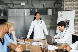 photo a team of health professionals seated at a table, one in a white lab coat is standing leading the group in discussions