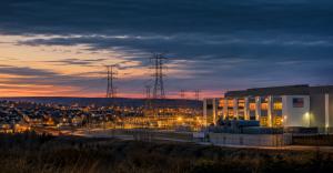 Cinematic editorial photograph showing modern American industrial landscape including data center facility, high-voltage transmission infrastructure, and residential community at twilight, representing the third major US housing era driven by industrial r