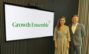 Growth Ensemble Co-founders and Managing Partners Meredith Carson and Adam Flinter standing next to a TV screen with their company logo on it.
