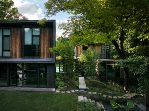 Native plantings in geometric blocks frame a modern home overlooking a wooded ravine, with a rustic plank bridge leading through a natural courtyard garden of steel, stone, and woodland-inspired materials.