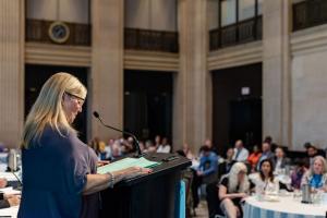 A woman stands on a podium talking to a room of attendees.