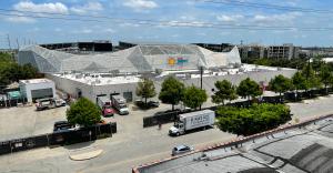 High angle photo of Shell Energy Stadium in EaDo, Houston, with nearby streets, trucks, trees, rooftops, and a blue sky with clouds.