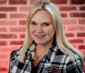 Professional portrait of a person wearing a Staff Zone branded plaid shirt, photographed against a red brick wall background.