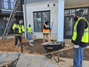 Four construction workers in high-visibility vests use tools and level soil outside a building under construction, with a wheelbarrow and ladder visible on site.