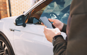 Person using a smartphone beside a used car, representing Car Owl vehicle history checks for UK drivers considering used electric cars.