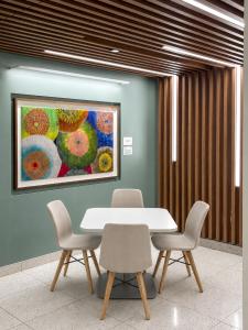 A small meeting nook at Scripps Encinitas featuring a white square table surrounded by four cream upholstered chairs with wooden legs, set against a sage green wall. A large framed artwork displaying colorful circular, radiating patterns in orange, green,