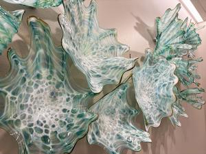 A close-up detail of a hand-blown glass wall sculpture at Scripps Encinitas. Several large, organic glass forms fill the frame, each shaped like ruffled, spiky sea creatures or coral with jagged, pointed edges. The glass is translucent and layered with sw