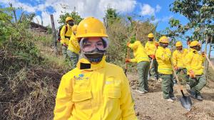 A group of women and men in yellow protective gear and helmets stand on a hillside carrying shovels.