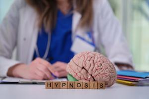 phot of a health professional in a lab coat sitting with a model of a brain with the words Hypnosis in front