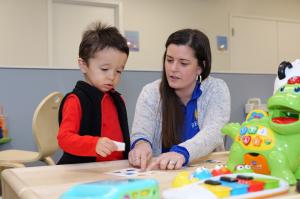 A Bierman Autism Centers clinician sits with a young boy at a therapy table, pointing to a card of colored shapes during an early learning activity.