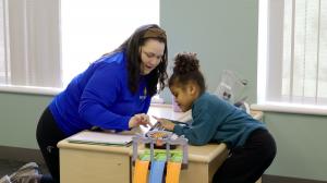 A Bierman Autism Centers clinician leans in beside a young child at a therapy table, guiding their hand toward a tablet during a play-based session.