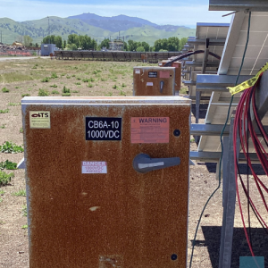  Close-up of a heavily rusted and weathered solar combiner box labeled "CB6A-10 1000VDC" with warning labels indicating high voltage. Red electrical cables are visible to the right, and solar panel racking is partially visible. A mountain range and constr