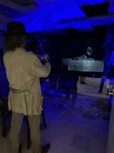 California Creative Corps / Culver City Artist Laureate Emeritus, Alexey Steele views the Ward That Never Closed at Hospital of Emotions with Alexey standing with an iPhone taking a picture of the IED exploded room with Holographic person